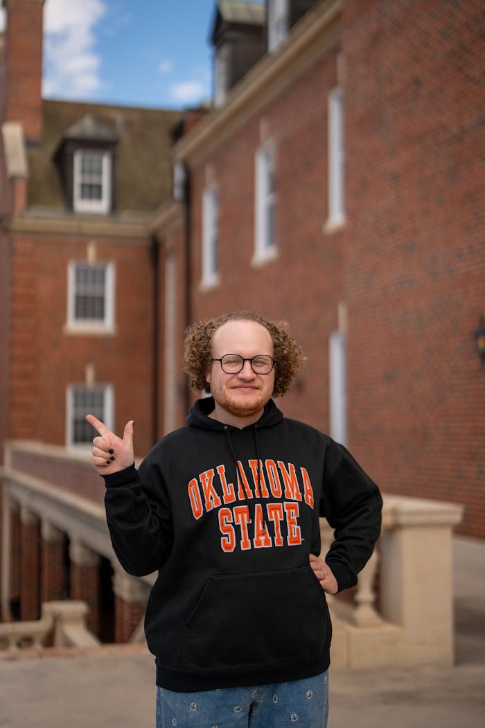 Young adult male wearing Oklahoma State hoodie standing on university campus.